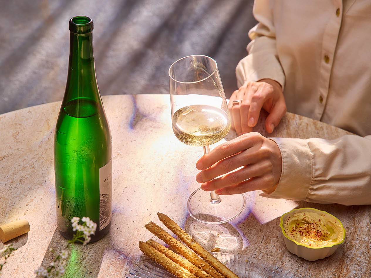 Eine Person hält ein Glas Weißwein, neben einer Flasche Wein und einem Teller mit Grissini und Hummus.