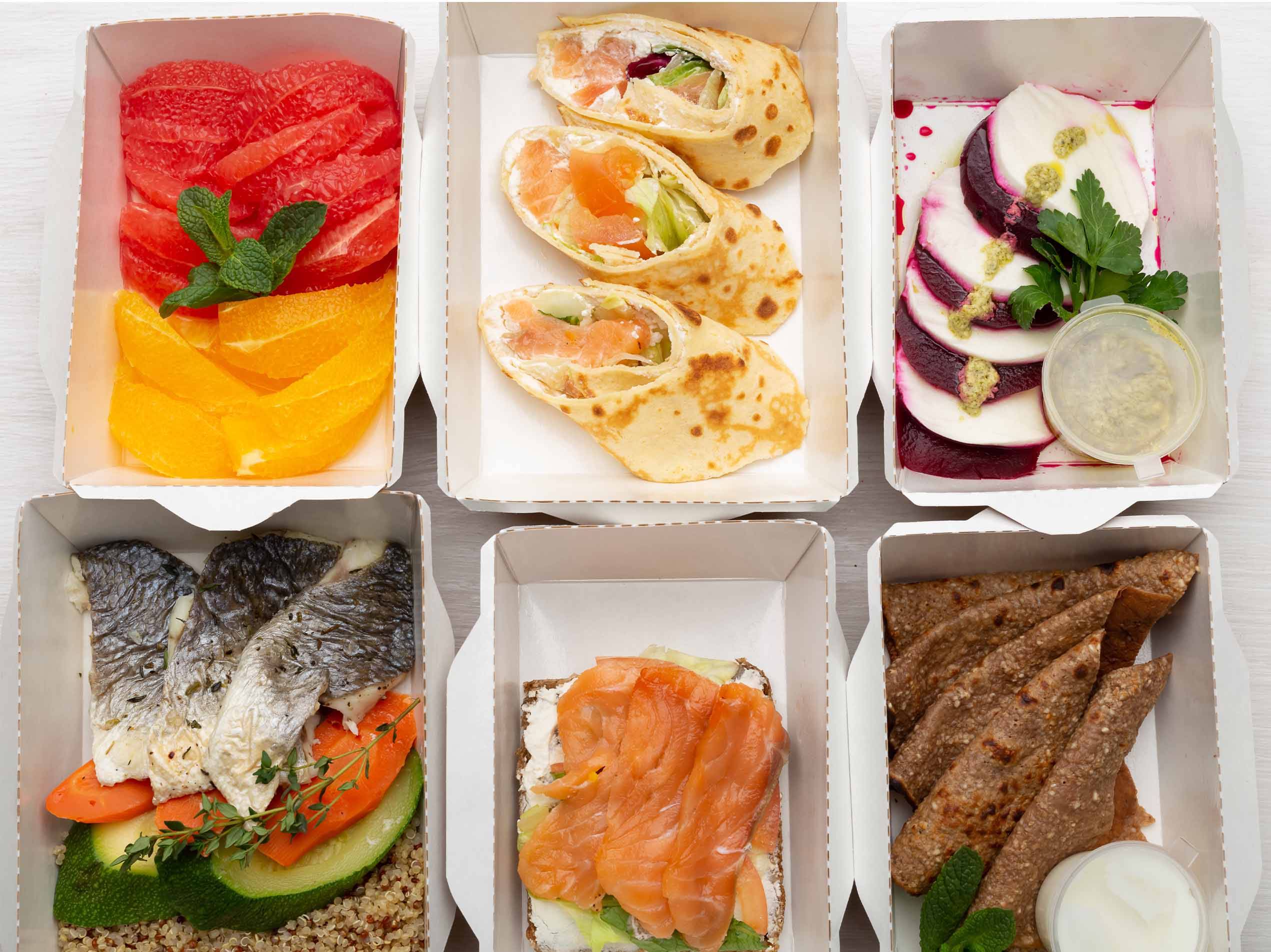 Sechs offene Essensboxen mit verschiedenen Mahlzeiten, darunter Obst, Wraps, Fisch und Brot.