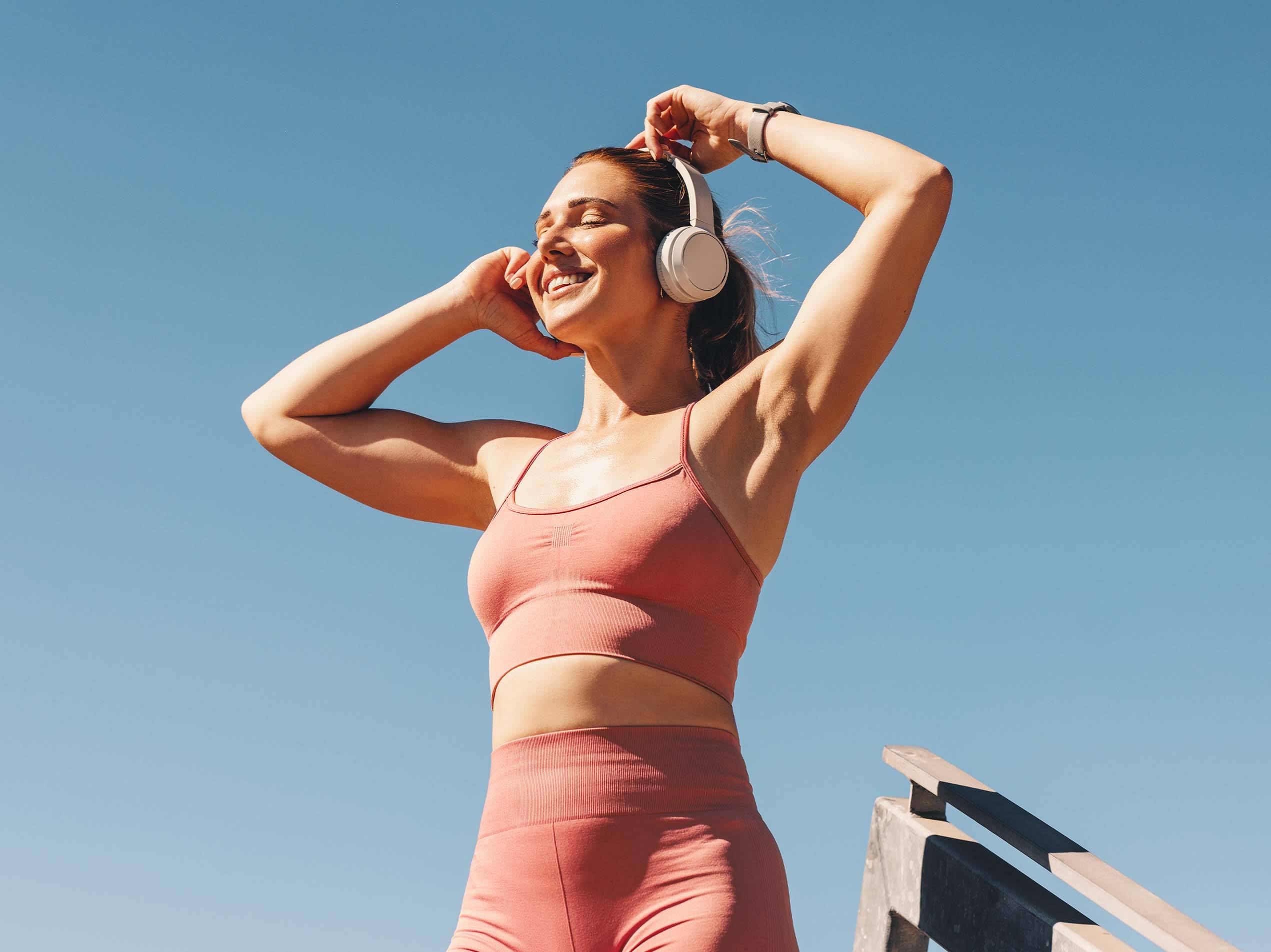 Lächelnde Frau in Sportkleidung mit Kopfhörern vor blauem Himmel.