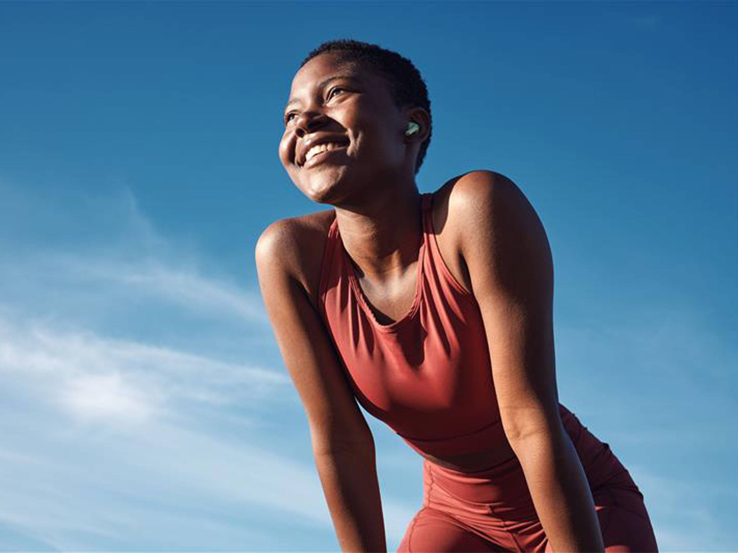 Eine lächelnde Frau mit In-Ear-Kopfhörern in Sportkleidung vor blauem Himmel.