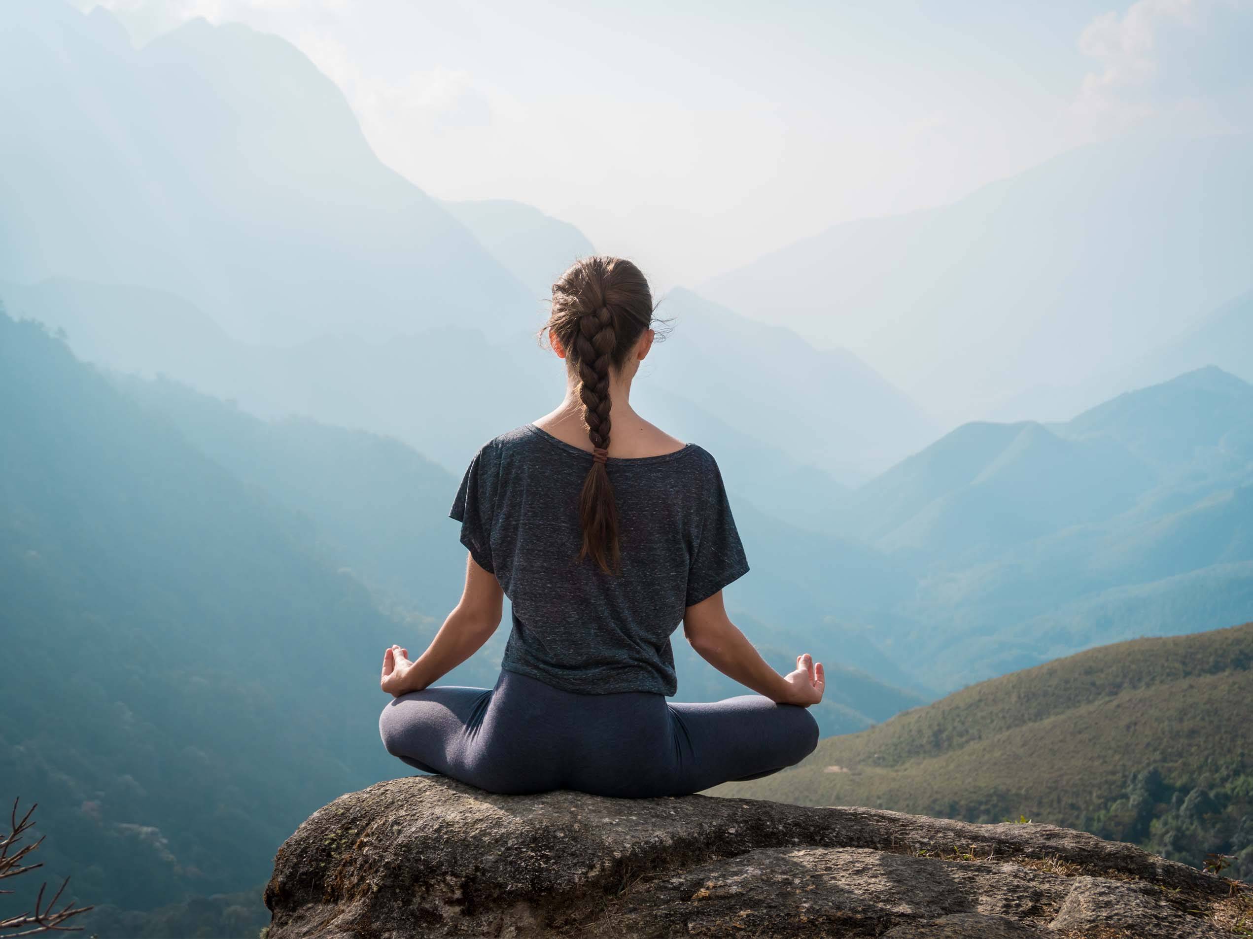 Eine Frau meditiert auf einem Felsen mit Blick auf eine Berglandschaft.