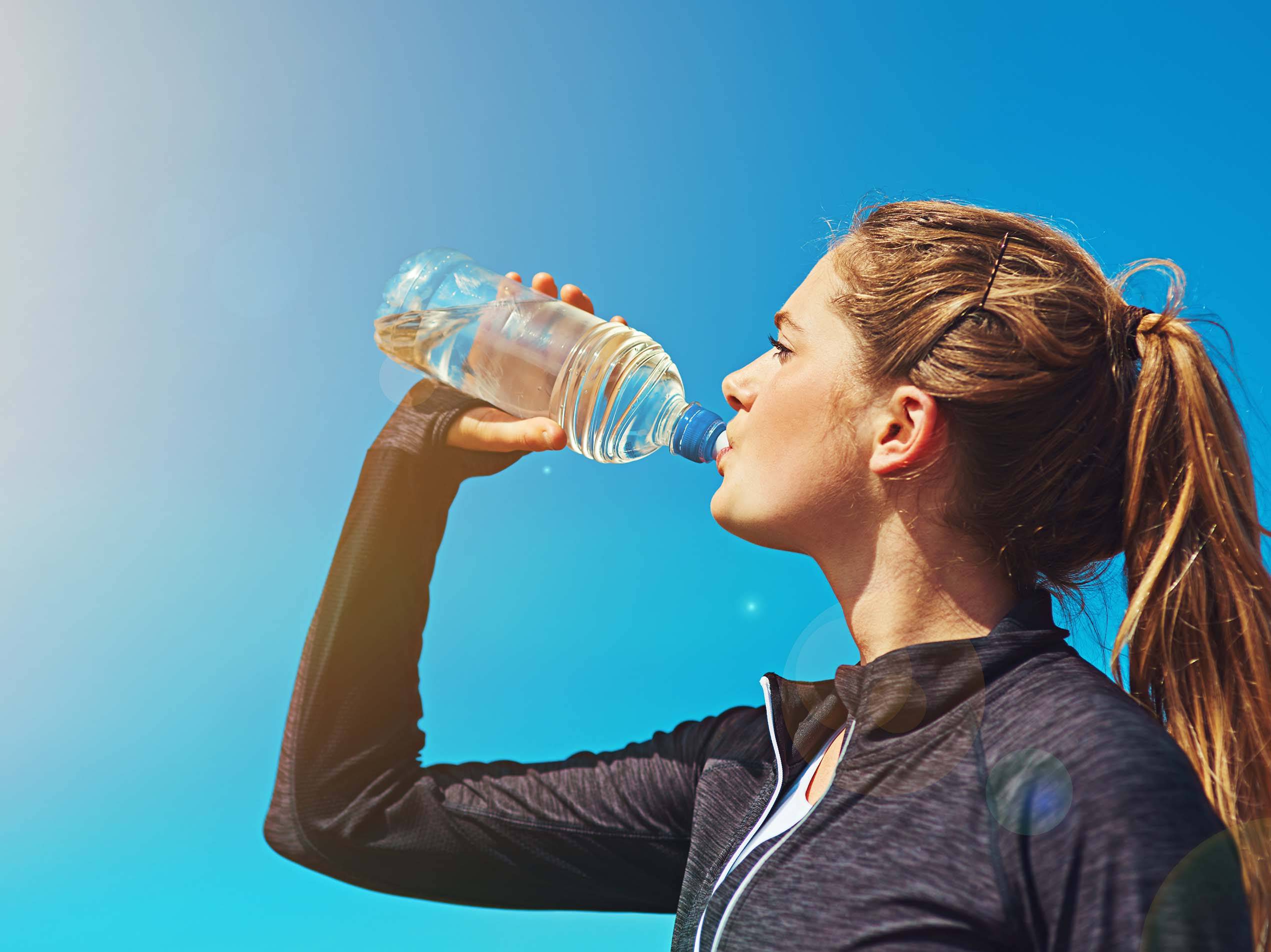 Junge Frau trinkt Wasser aus einer Plastikflasche vor blauem Himmel.