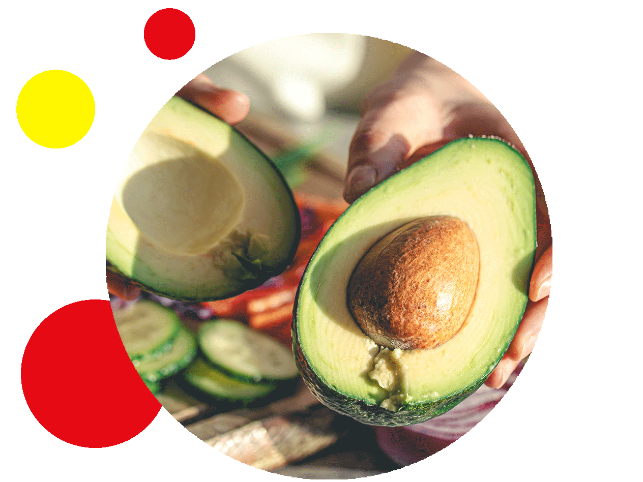 Hände halten eine aufgeschnittene Avocado, daneben geschnittene Gurken.