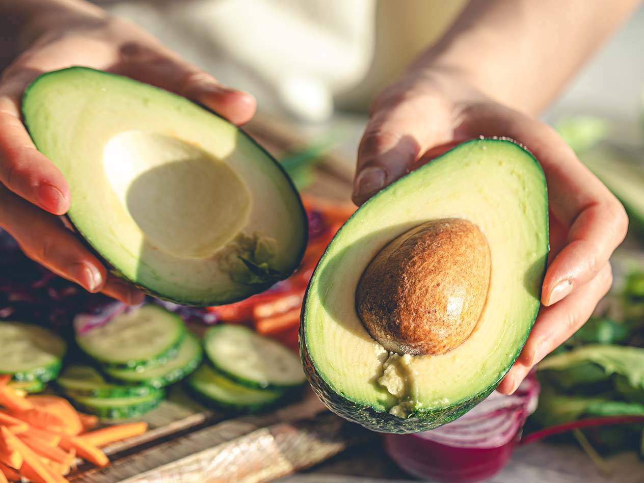 Hände halten eine halbierte Avocado über geschnittenem Gemüse.
