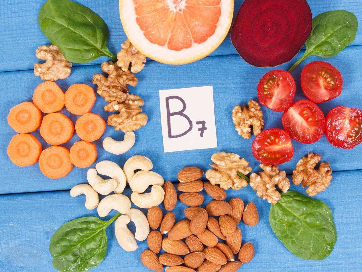 Eine Auswahl an Lebensmitteln, die reich an Vitamin B7 sind, darunter Nüsse, Gemüse und Obst, auf blauem Hintergrund.