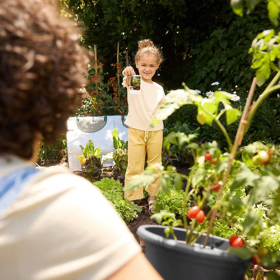 Kind im Garten hält ein Foto, mit frischen Produkten und einer Tomatenpflanze im Vordergrund.
