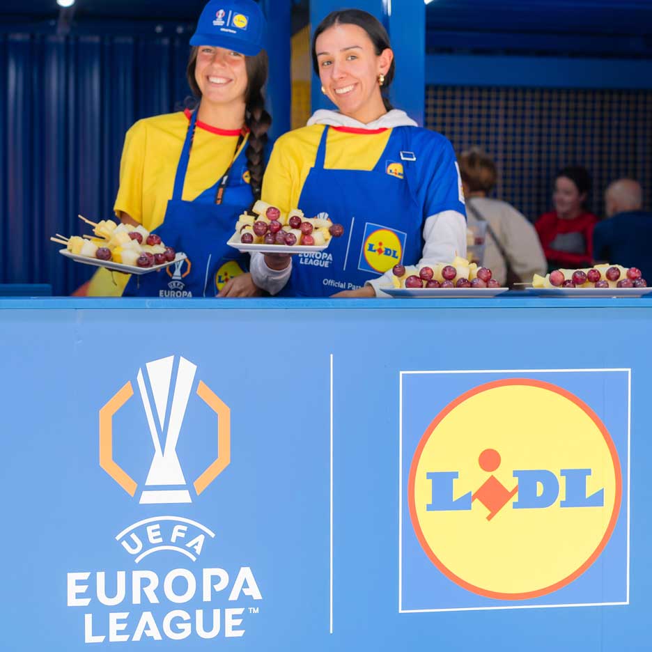 Zwei lächelnde Frauen in Lidl-Schürzen bieten Obstspieße bei einem UEFA Europa League-Event an.