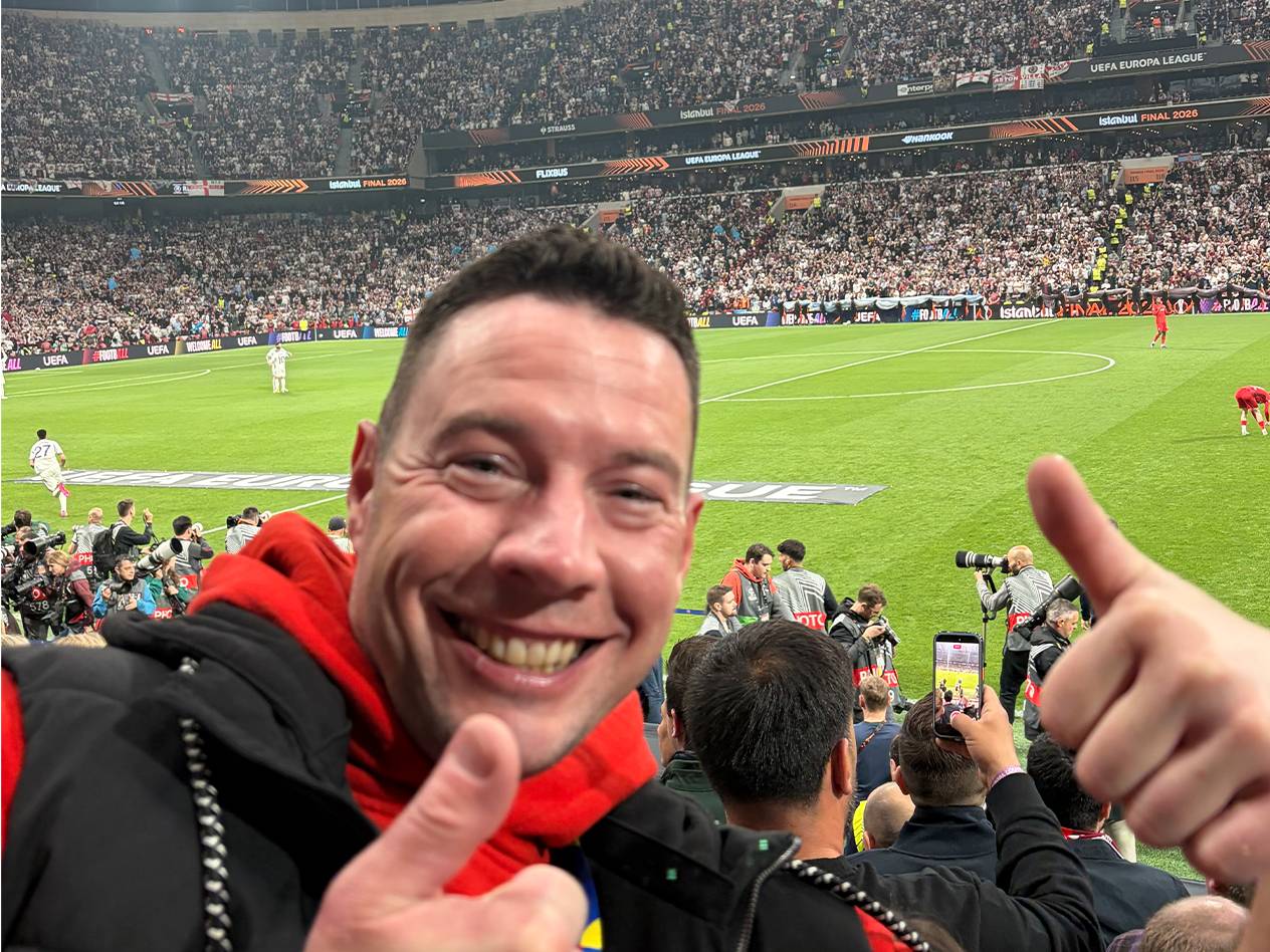 Ein lächelnder Mann im roten Hoodie gibt im Stadion zwei Daumen hoch, im Hintergrund ein Fußballspiel.