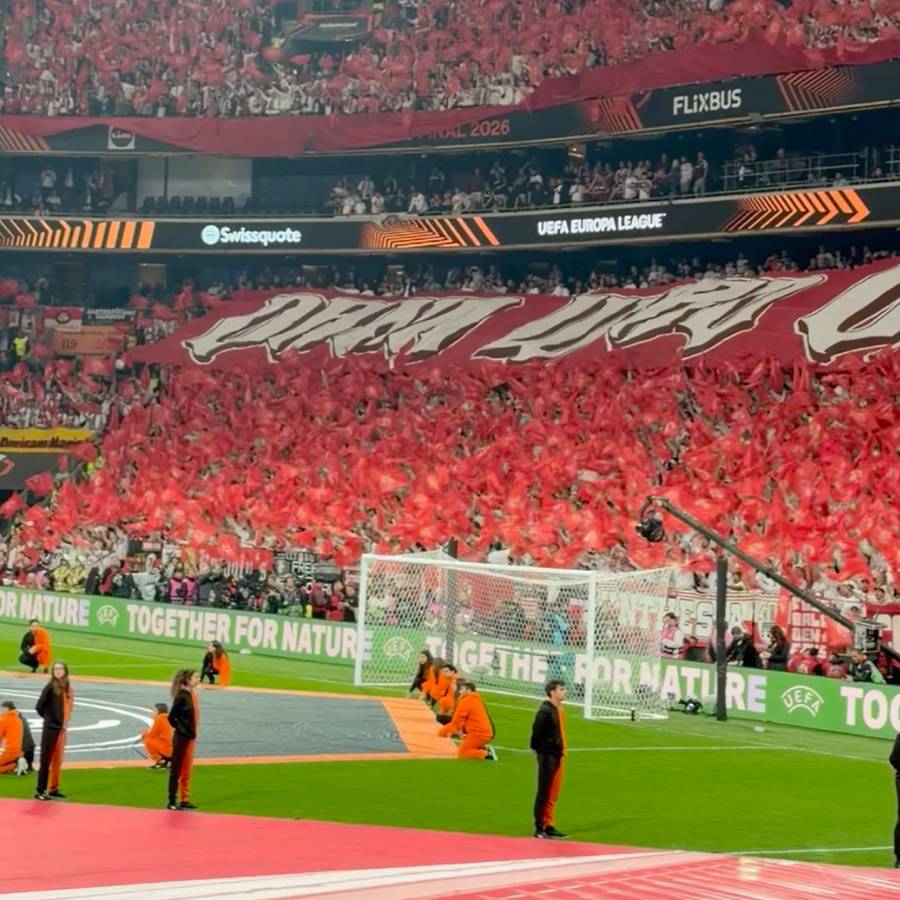 Fußballstadion mit Fans, die rote Fahnen schwenken, und einem Banner mit der Aufschrift „TOGETHER FOR NATURE“.