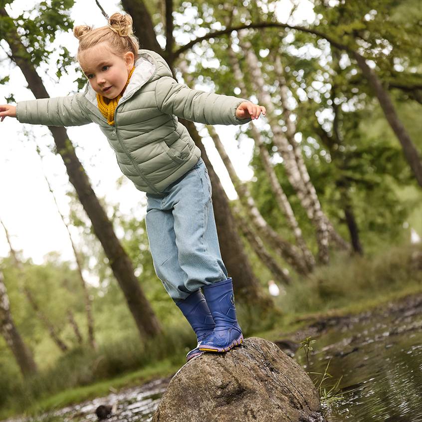 Kleines Mädchen in Steppjacke, Jeans und Gummistiefeln balanciert auf einem Felsen im Wald.