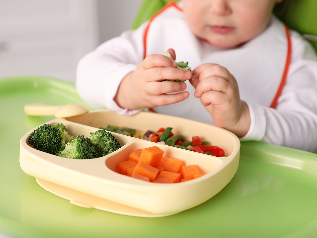 Baby in Hochstuhl mit geteiltem Teller voller Gemüse