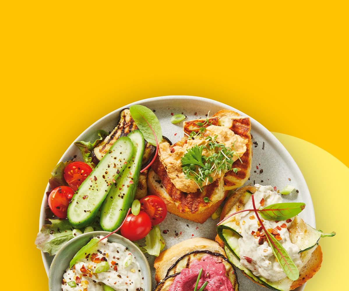 Teller mit Bruschetta, Hummus, gegrilltem Gemüse, Gurken und Tomaten auf gelbem Hintergrund.