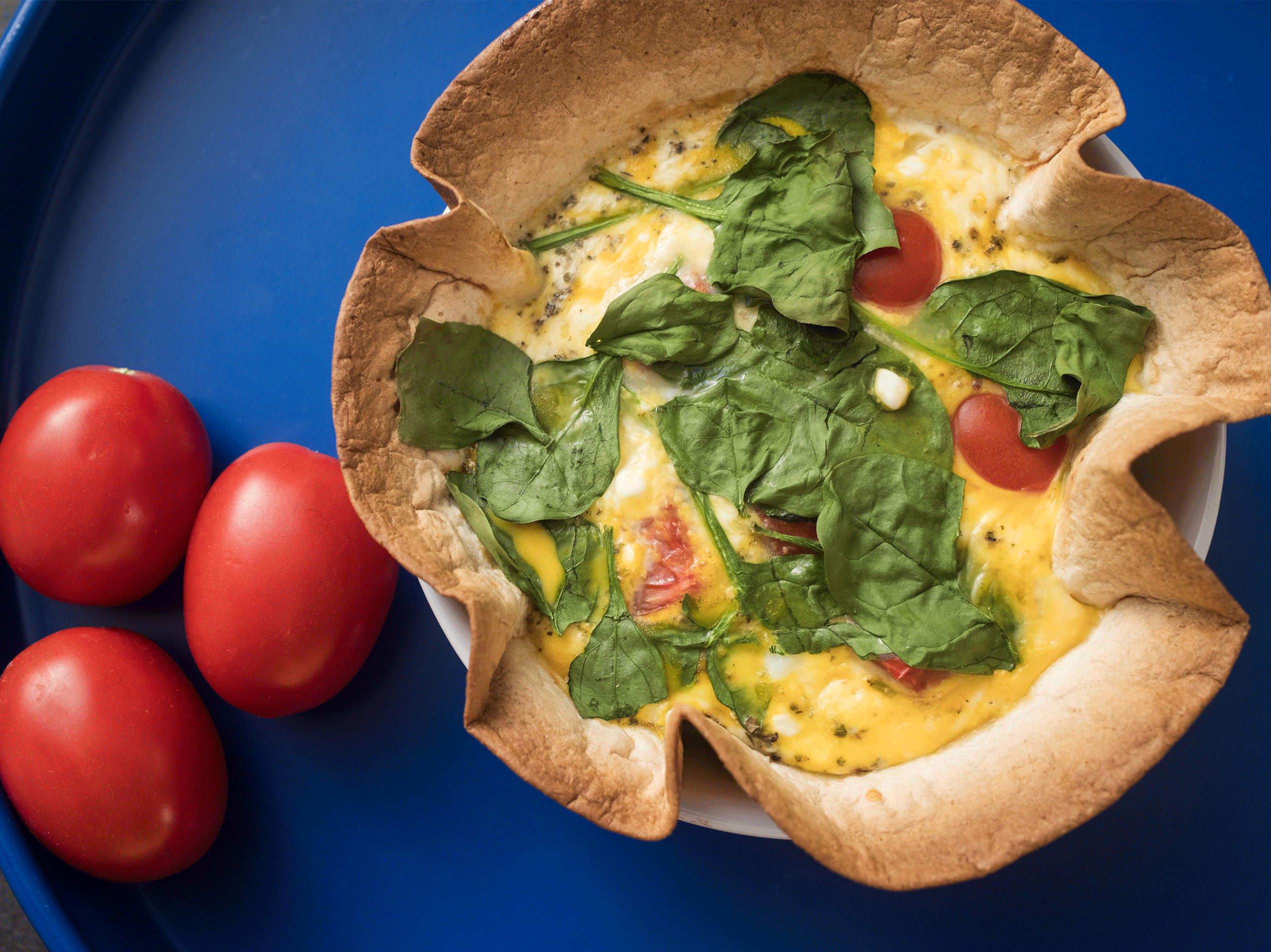Quiche mit Spinat und Tomaten in Tortilla-Schale auf blauem Tablett mit frischen Tomaten.