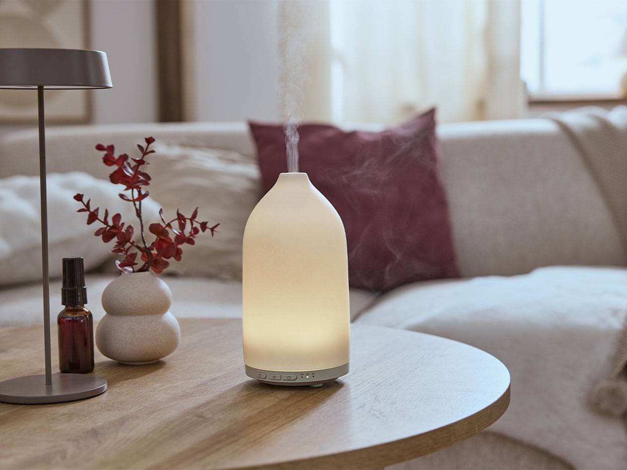 Beleuchteter Aroma-Diffusor, Ölflasche und Vase mit Zweigen auf einem Tisch.