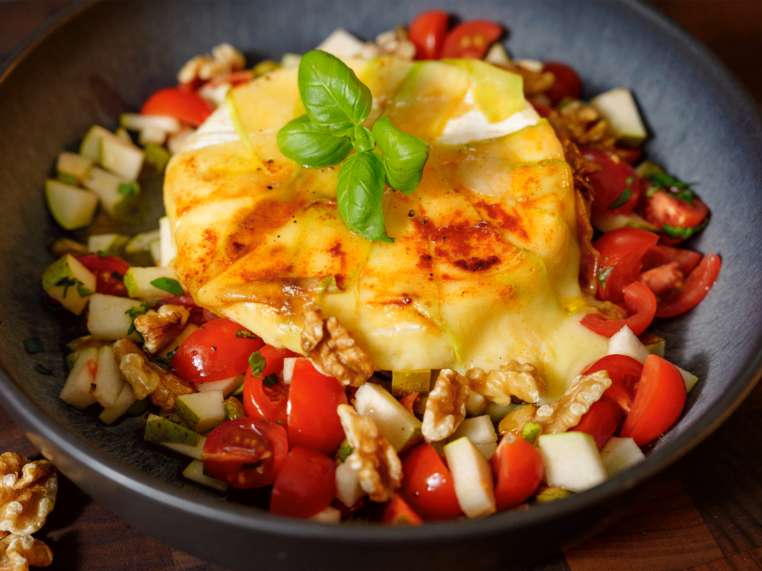 Gebackener Camembert mit Birnen-Tomaten-Salat, Walnüssen und Basilikum in einer dunklen Schale.