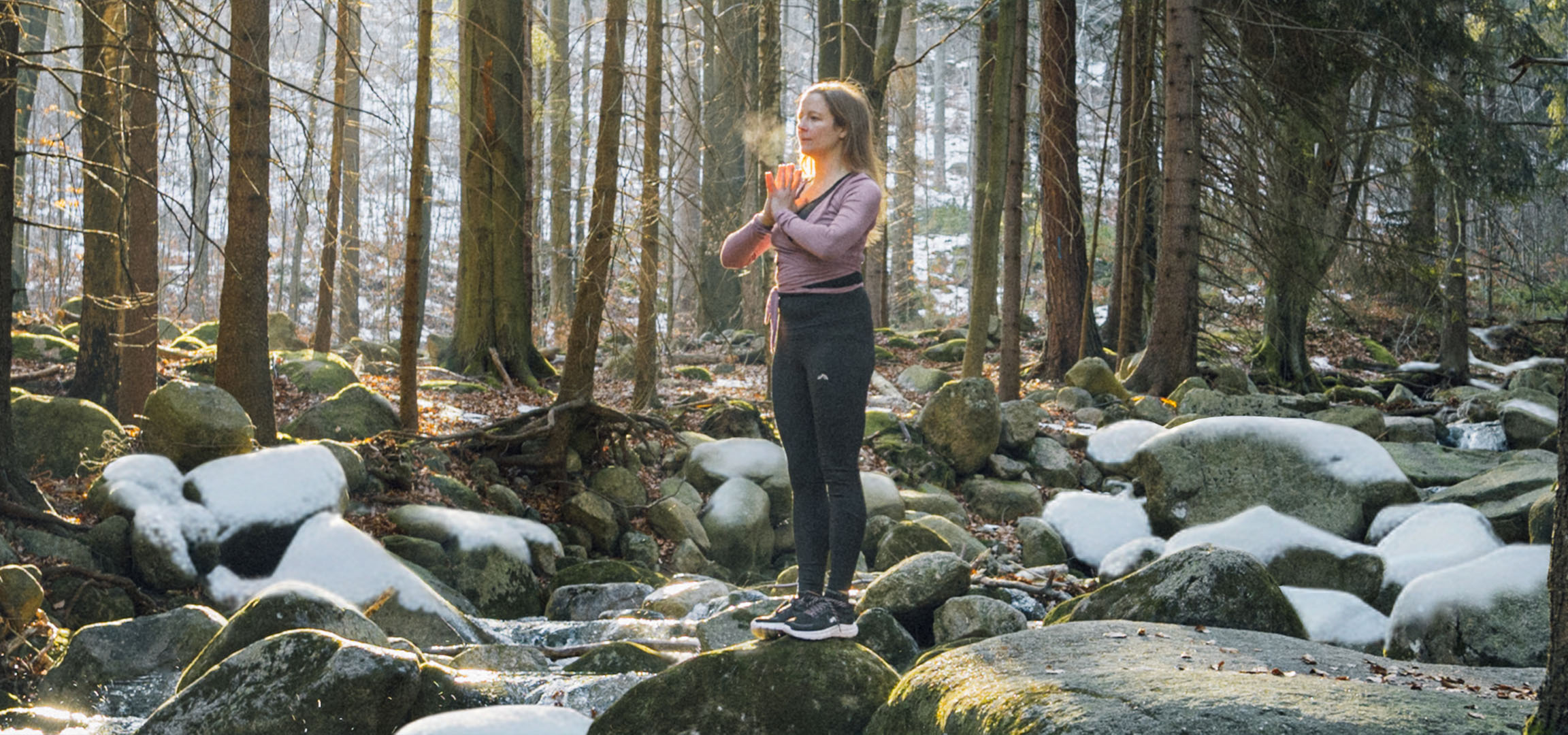 Frau in Sportkleidung macht Yoga in einem verschneiten Wald mit Bach.