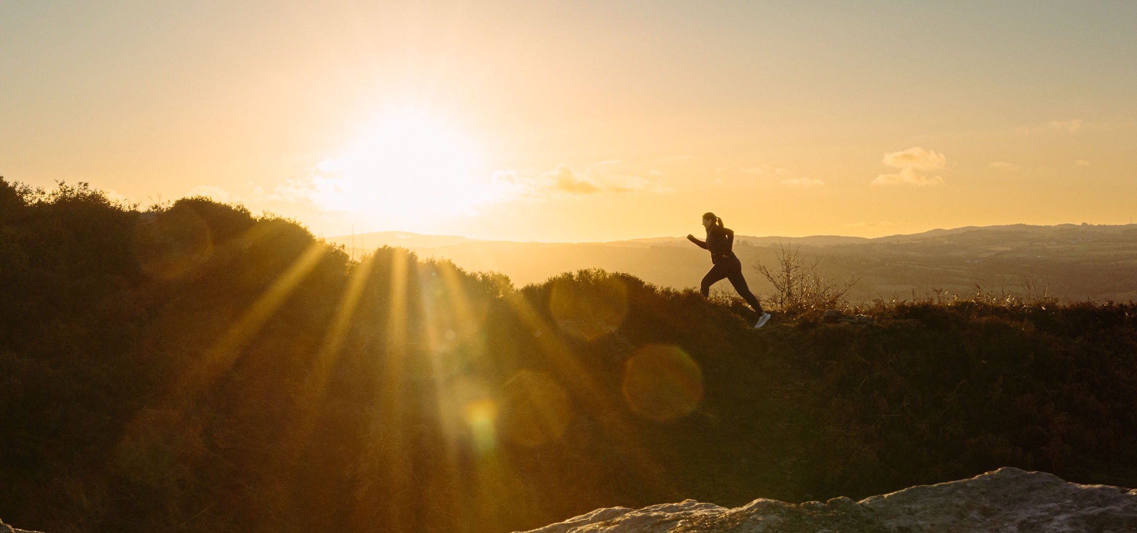 Frau läuft bei Sonnenuntergang auf einem Hügel, Sonnenstrahlen scheinen durch die Landschaft.