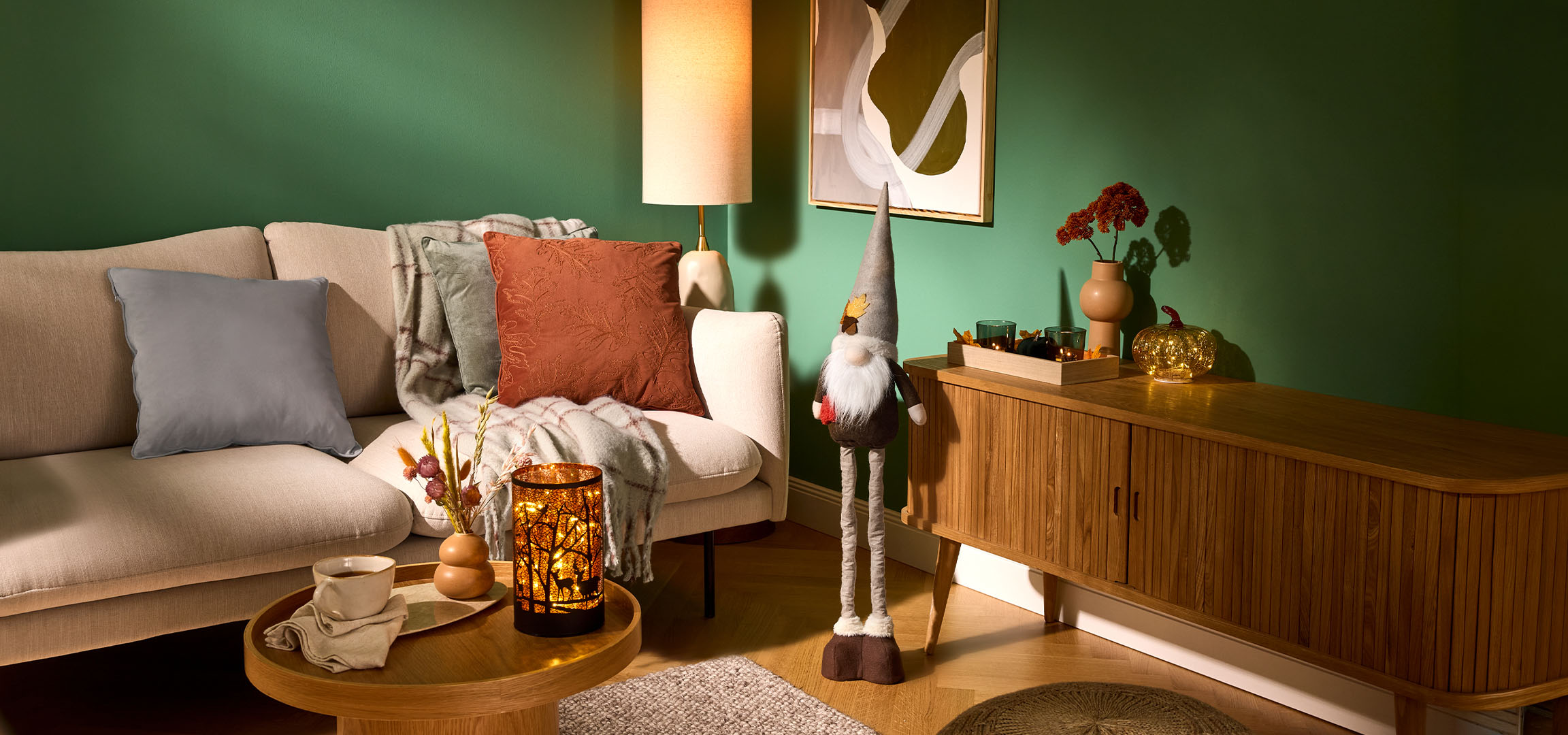 Gemütliches Wohnzimmer mit beigem Sofa, Kissen, Decke und Herbstdekorationen.
