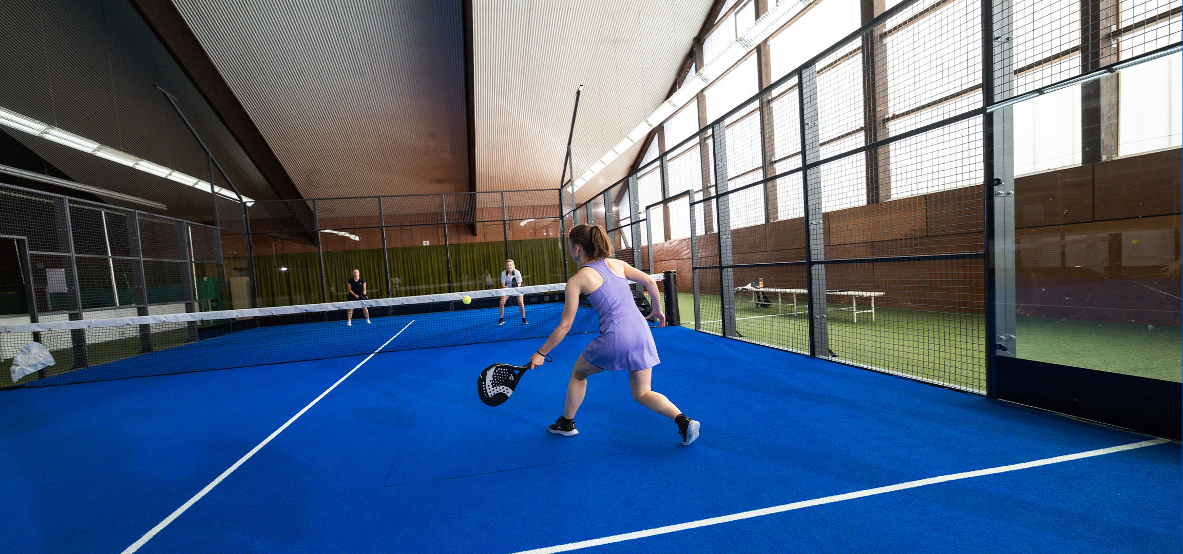 Frau spielt Padel auf einem blauen Platz, mit anderen Spielern im Hintergrund.