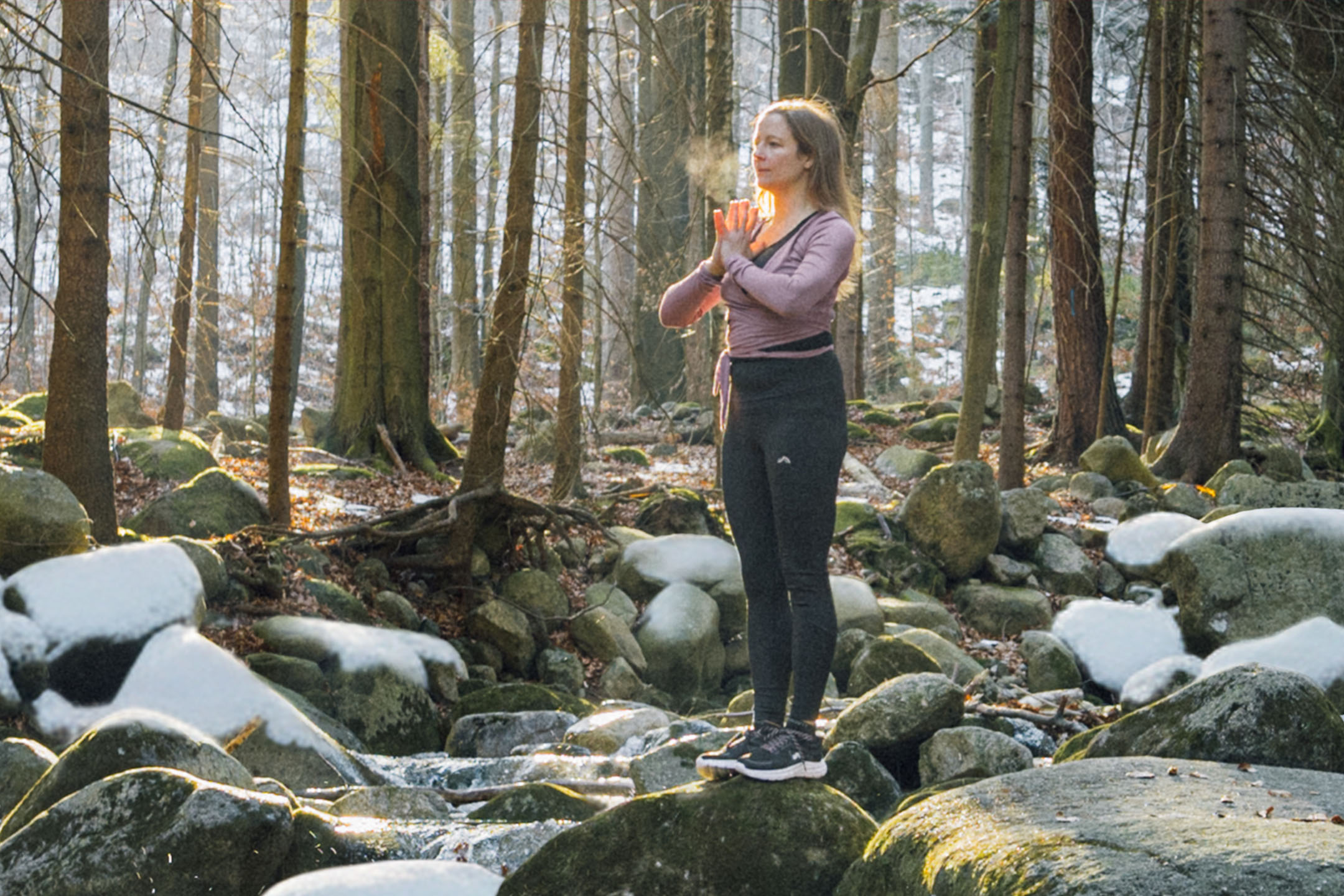 Frau in Sportkleidung praktiziert Yoga in einem Winterwald an einem Bach.