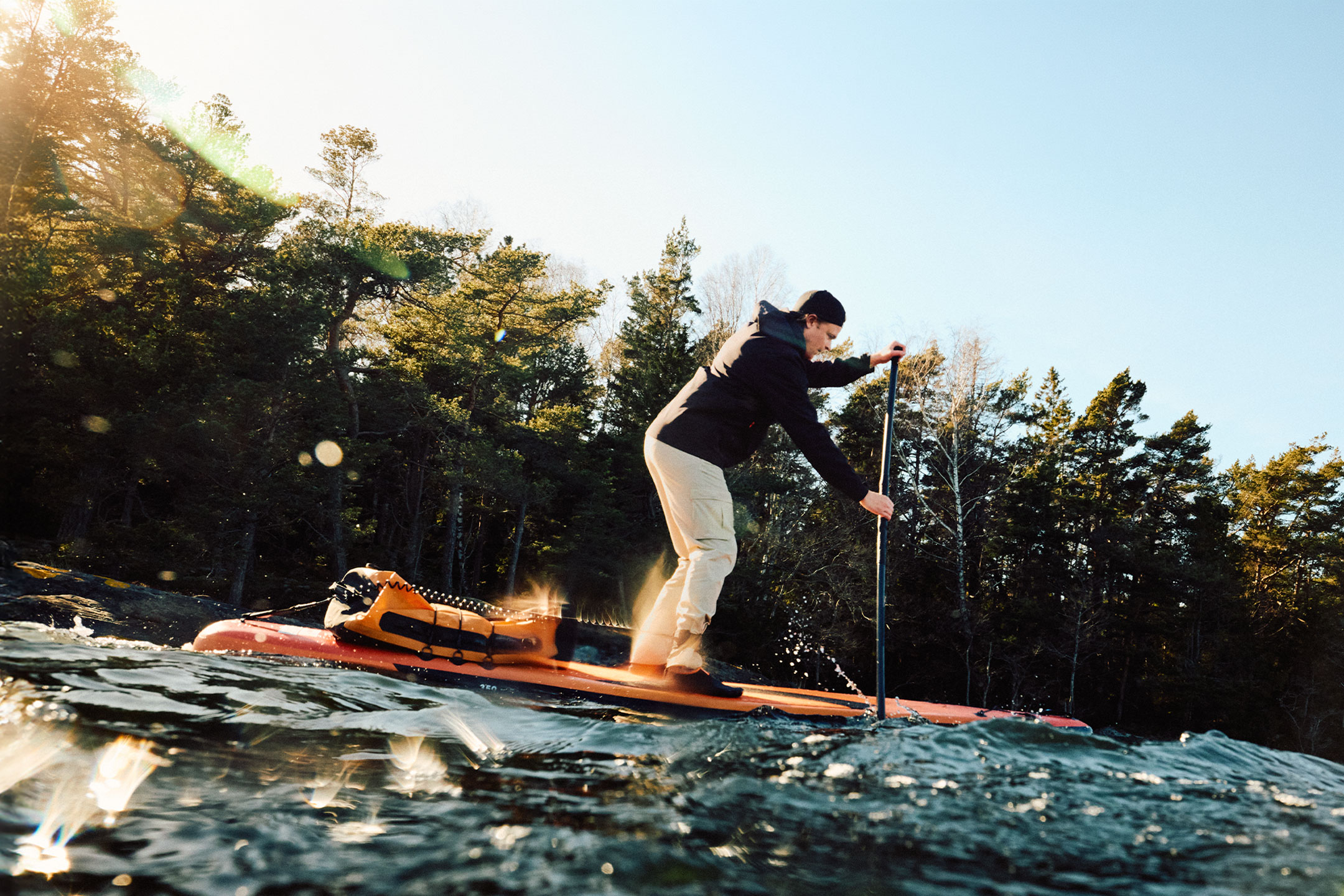 Mann paddelt auf einem Stand-Up-Paddleboard mit orangefarbenem Packsack am Waldrand.