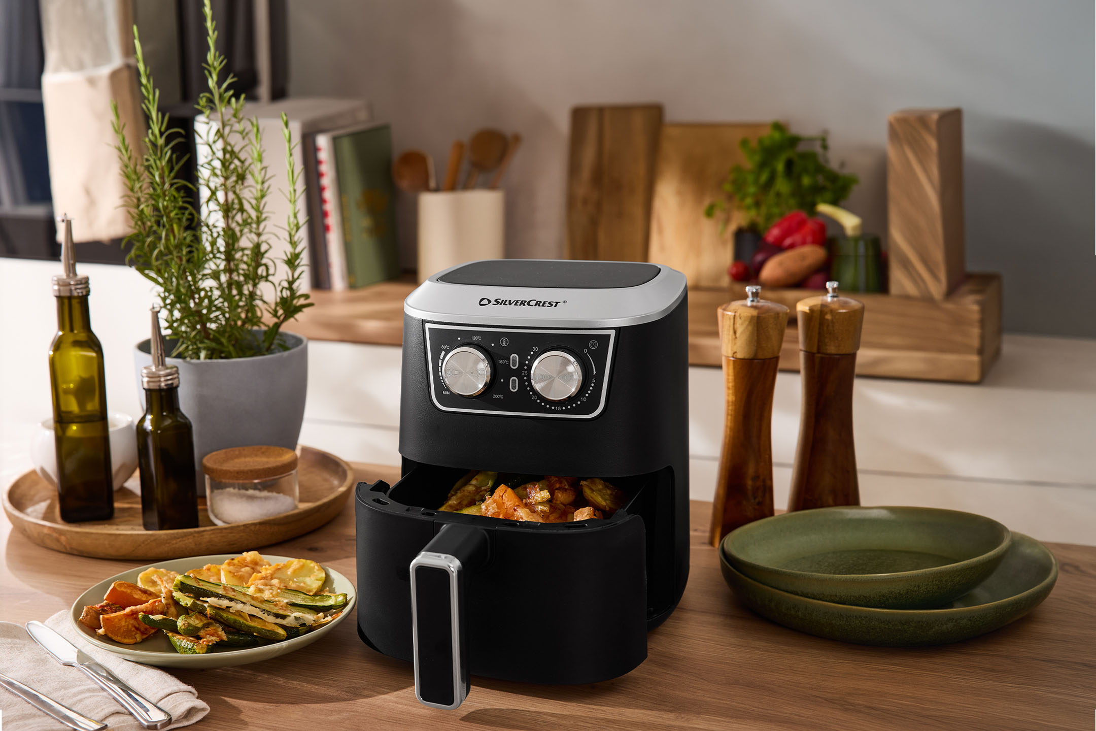 Schwarze Heißluftfritteuse mit Gemüse, Gewürzen und Geschirr auf einer Holztheke.