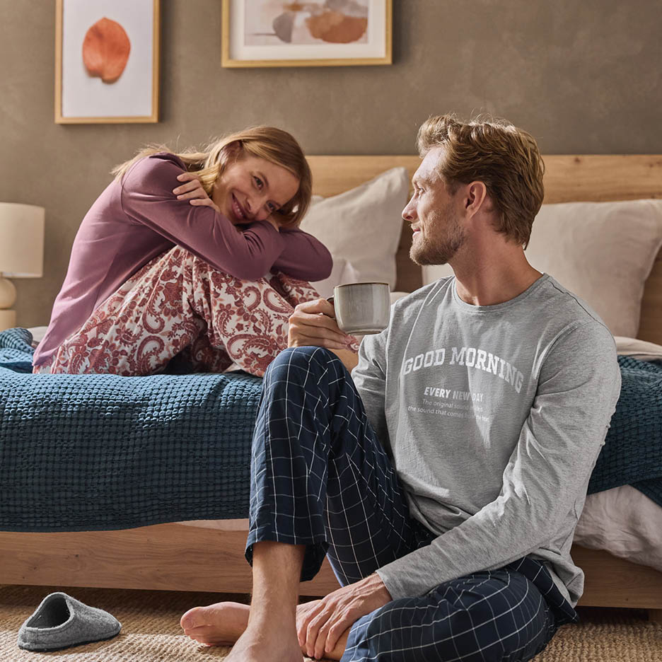 Ein Paar in Pyjamas sitzt im Schlafzimmer, der Mann trinkt Kaffee.