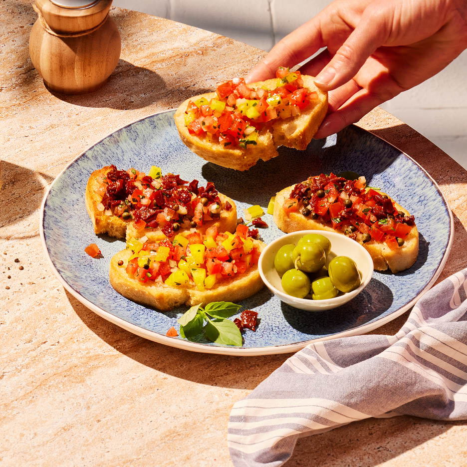 Eine Hand nimmt eine Bruschetta mit Tomaten und Paprika von einem Teller mit weiteren Bruschetta und Oliven.