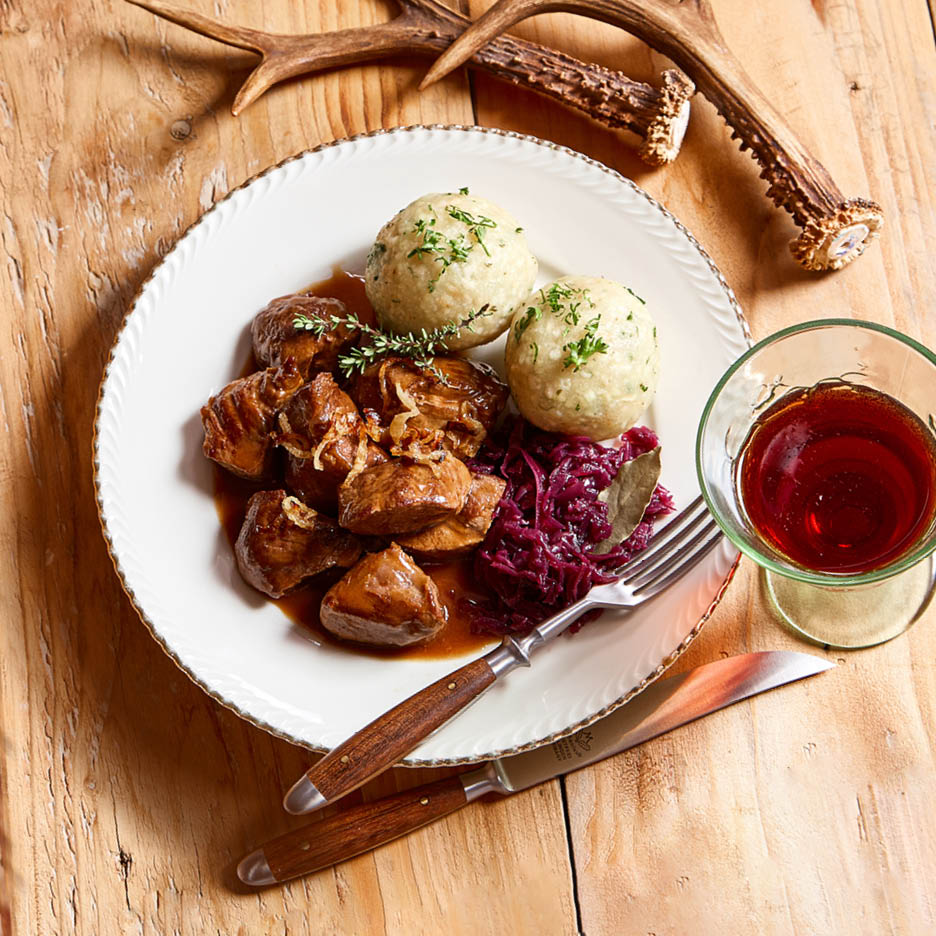 Ein Teller mit Gulasch, Knödeln und Rotkohl, daneben ein Glas Rotwein und Hirschgeweihe.