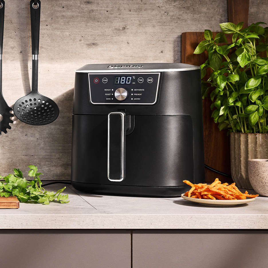 Ein schwarzer Airfryer von Silvercrest auf einer Küchenarbeitsplatte mit Pommes und Kräutern.