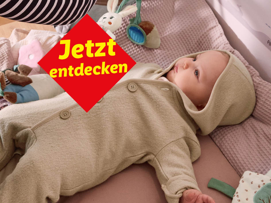 Ein Baby in einem beigen Strampler liegt in einem Kinderbett mit Spielzeug und der Aufschrift „Jetzt entdecken“.