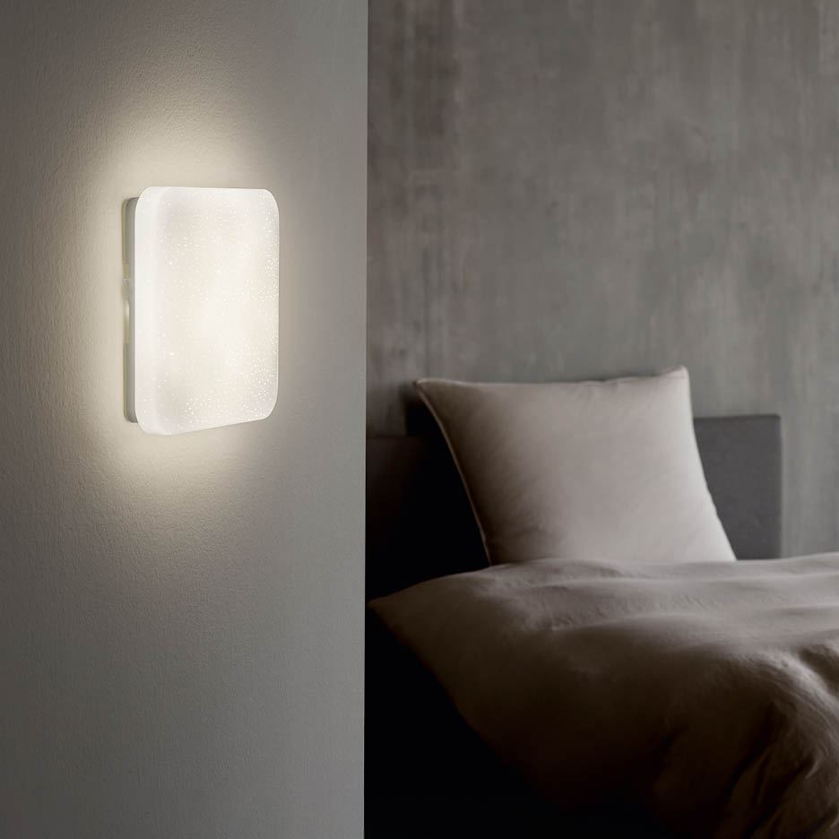 Beleuchtete Wandleuchte neben einem Bett mit Kissen und Decke.