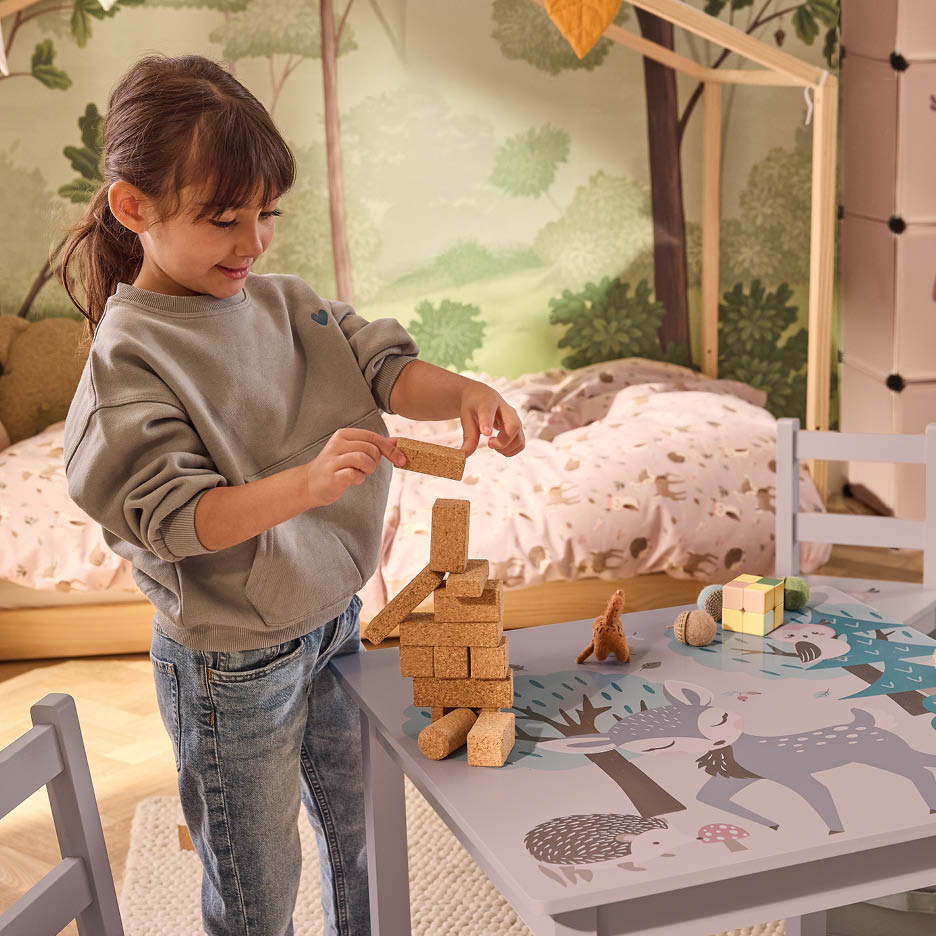 Ein Mädchen spielt mit Holzklötzen an einem Kindertisch mit Waldmotiv in einem Kinderzimmer.