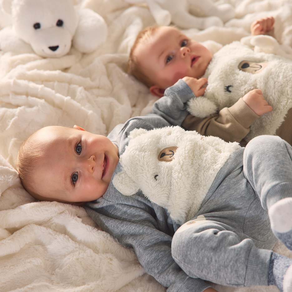 Zwei Babys in grauen Bären-Overalls liegen auf einer weißen Decke.