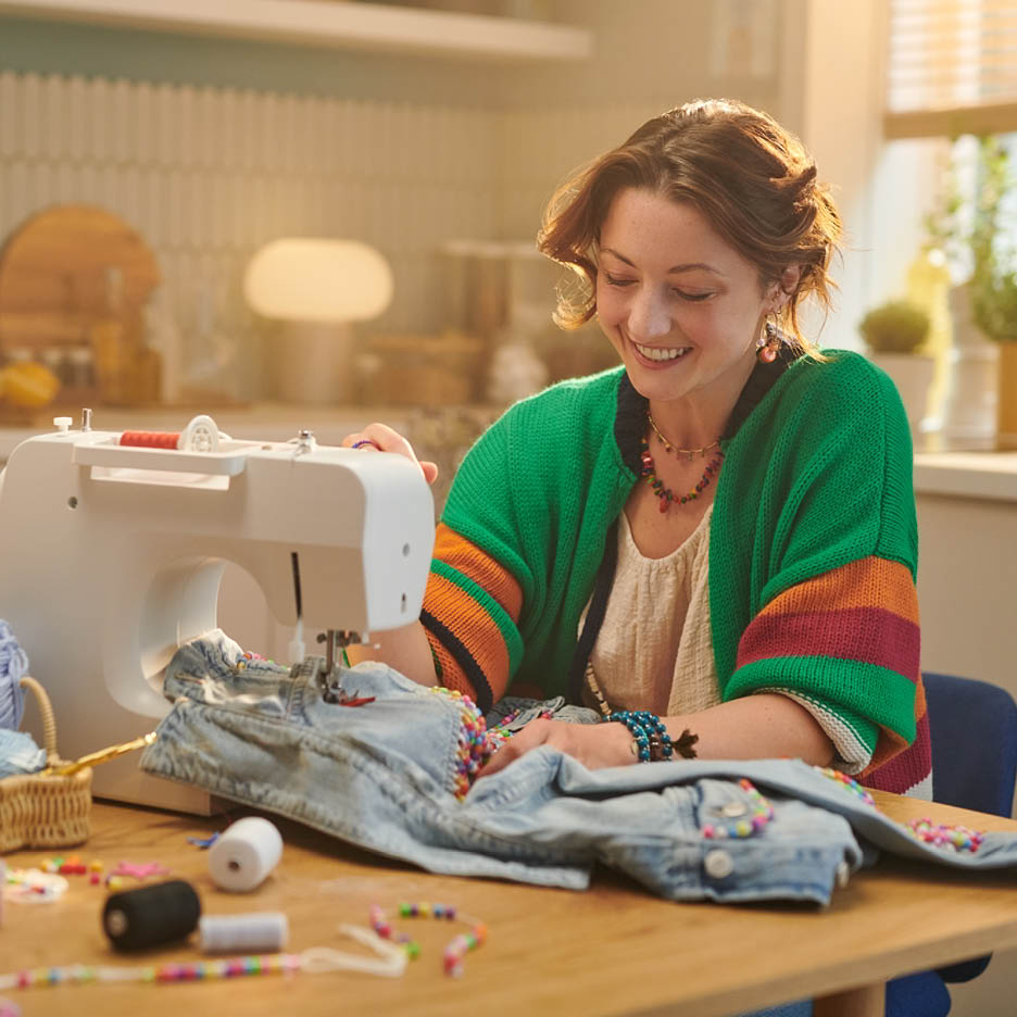 Eine lächelnde Frau näht Perlen auf Jeans mit einer Nähmaschine.