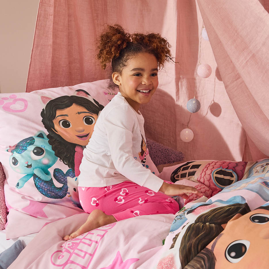 Ein lächelndes Mädchen in rosa Pyjama sitzt auf einem Bett mit Gabby's Dollhouse Bettwäsche.