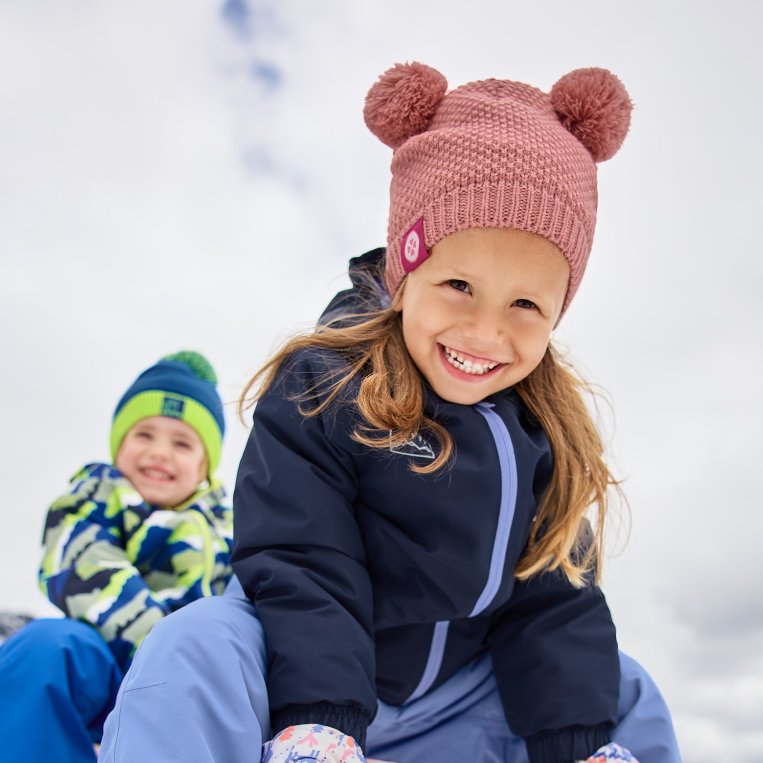 Zwei Kinder in Winterkleidung, das Mädchen trägt eine rosa Mütze mit Bommeln.