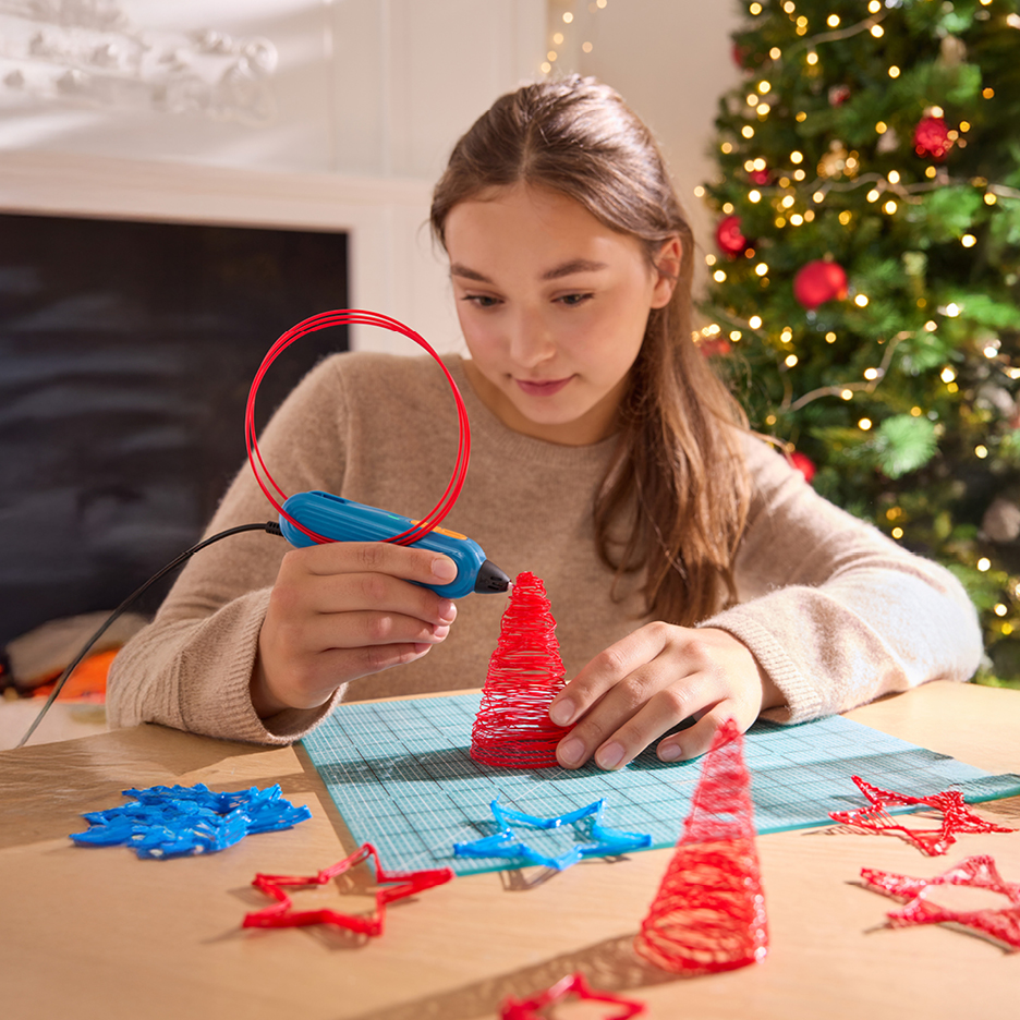 Mädchen bastelt mit 3D-Stift rote Weihnachtsdekorationen, darunter Bäume und Sterne.