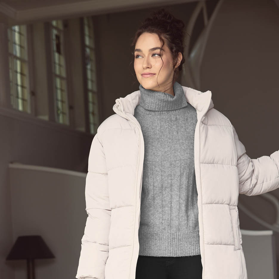 Eine Frau in einem beigefarbenen Pufferjacke und einem grauen Rollkragenpullover blickt nach links.