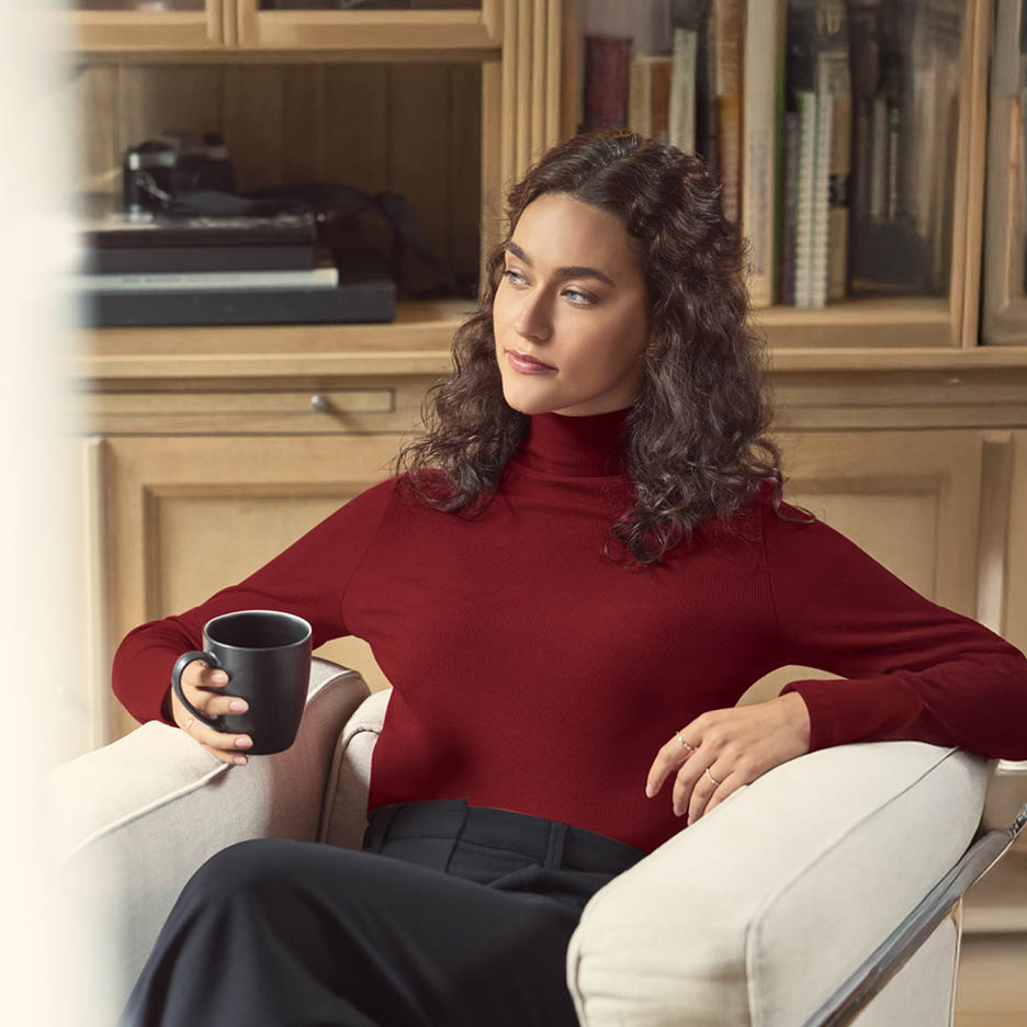 Frau in rotem Rollkragenpullover und schwarzer Hose sitzt mit einer Tasse in einem Sessel.