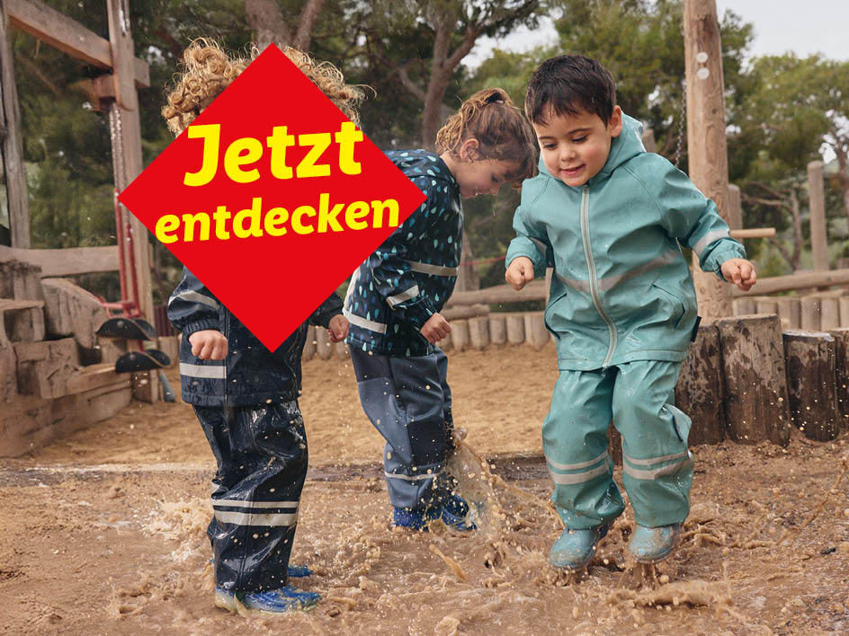Drei Kinder in Matschhosen und Regenjacken springen in Pfützen.