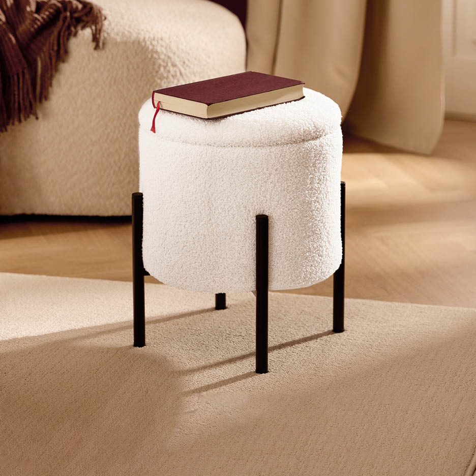 Ein cremefarbener Bouclé-Hocker mit schwarzen Beinen und einem Buch darauf, auf einem Teppich.
