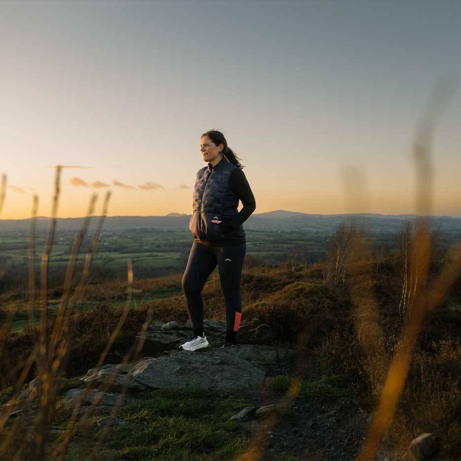 Frau in Steppweste und Leggings steht auf einem Hügel bei Sonnenuntergang.