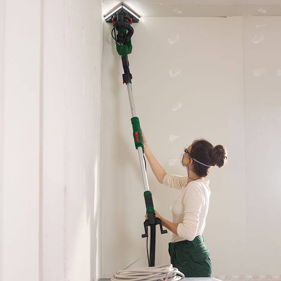 Frau schleift eine Wand mit einem Teleskop-Wandschleifer in einer Ecke.
