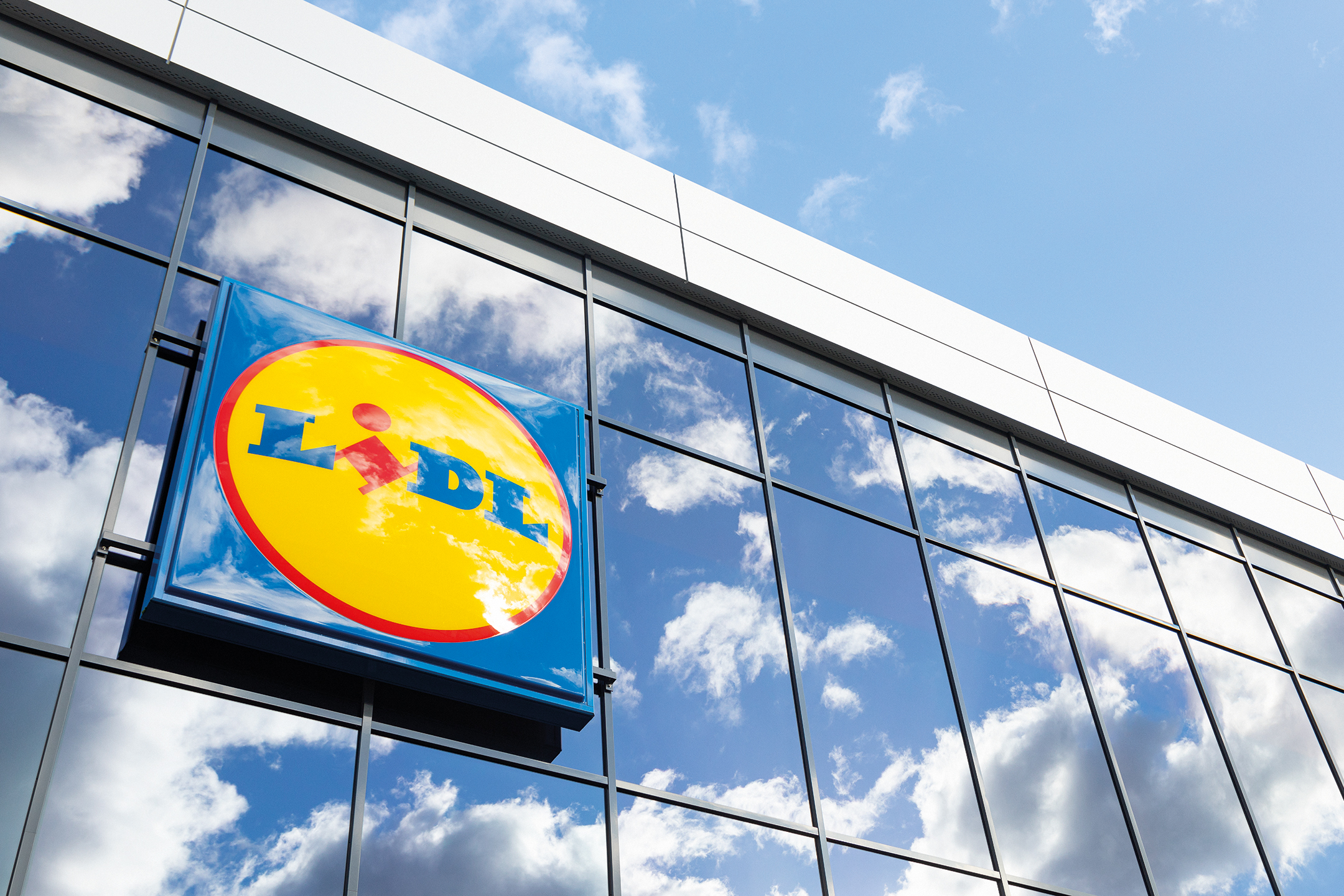 Lidl-Schild an einer Glasfassade mit Spiegelung von Himmel und Wolken.
