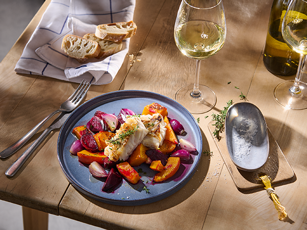 Gebratener Fisch mit Gemüse, Brot und Weißwein auf einem Holztisch.