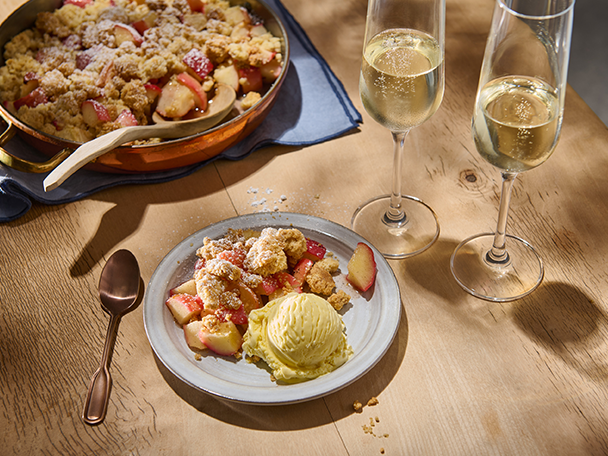 Apfel-Crumble mit Vanilleeis und zwei Gläsern Sekt auf einem Holztisch.