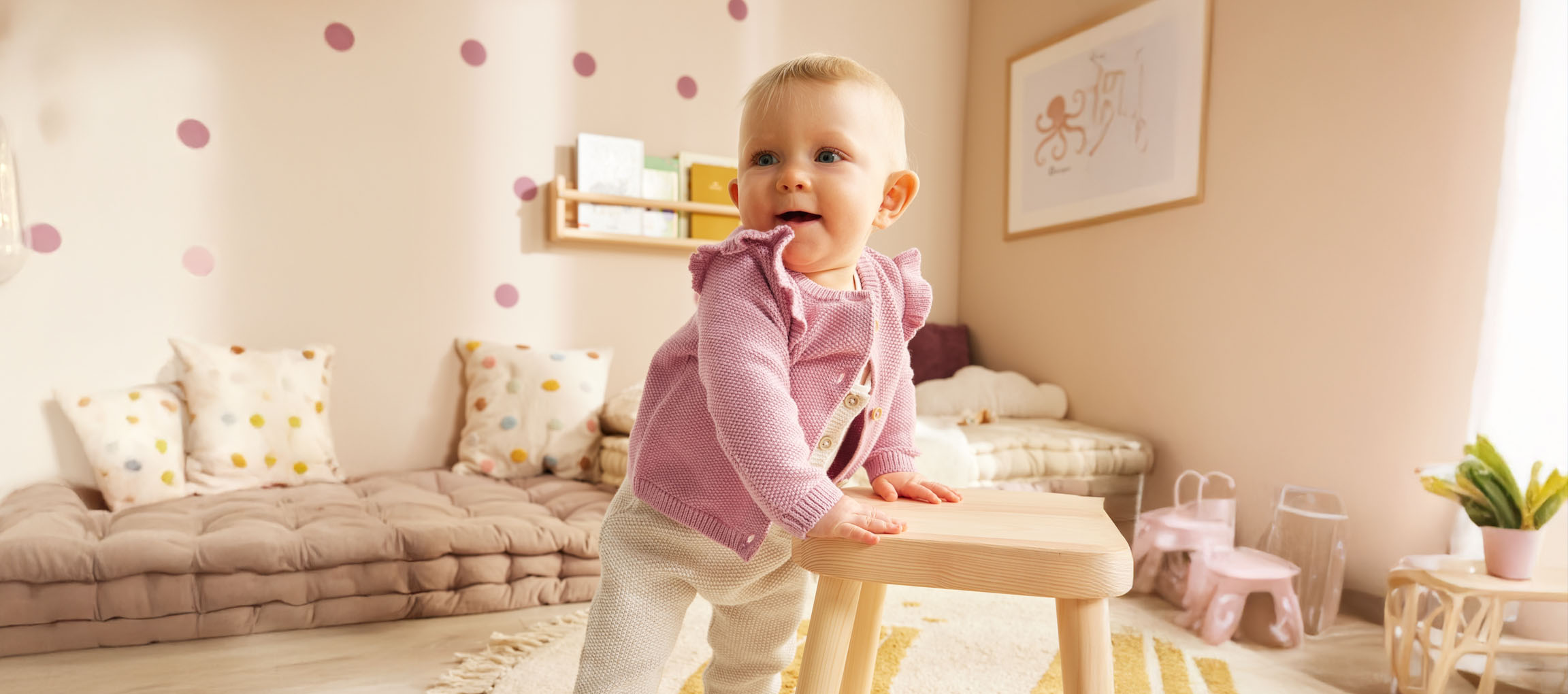Ein Baby in einem süßen rosa Cardigan steht neben einem Holzhocker in seinem Kinderzimmer.