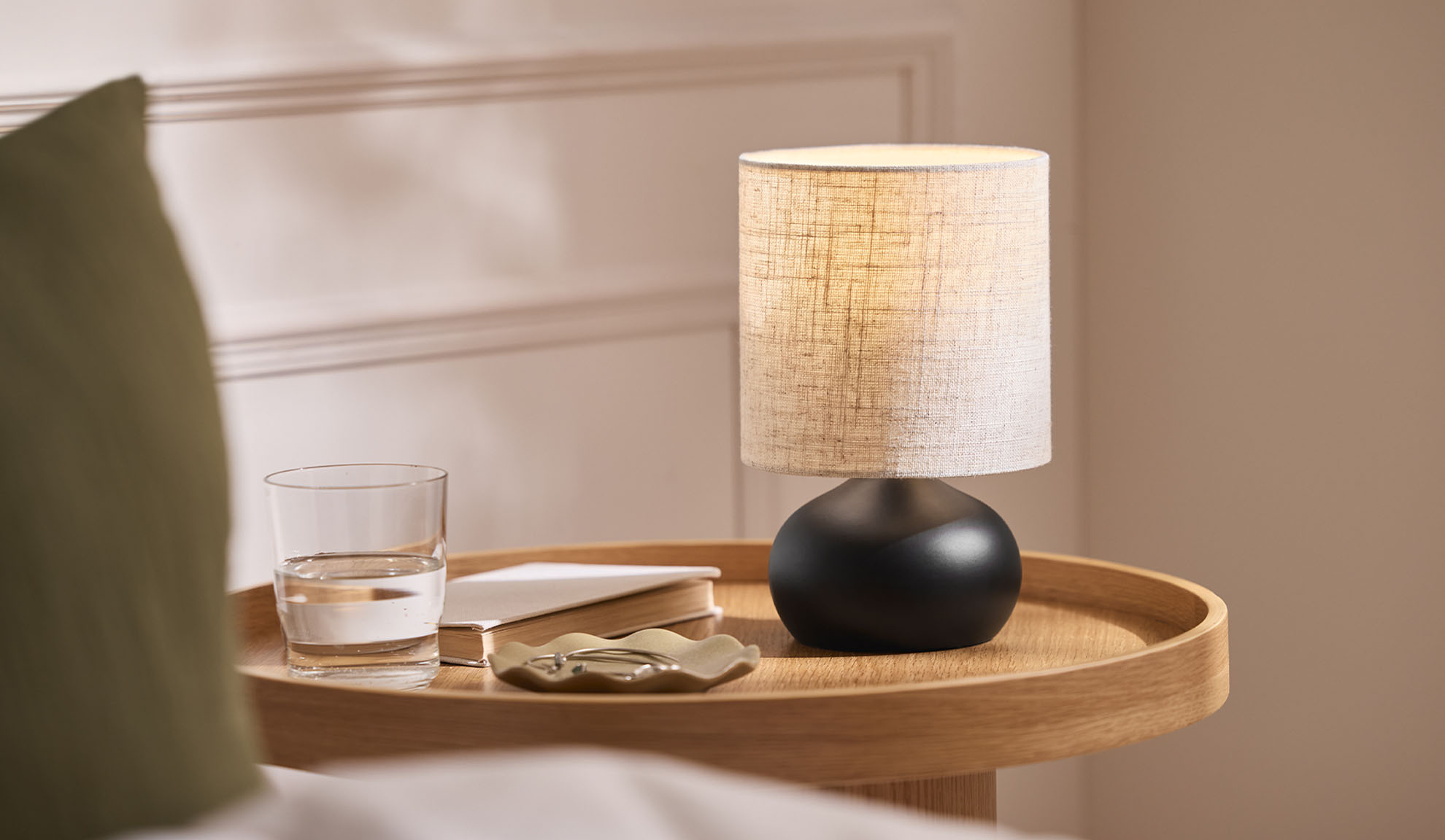 Nachttischlampe, Glas, Buch und Schmuckschale auf einem Holztablett neben dem Bett.