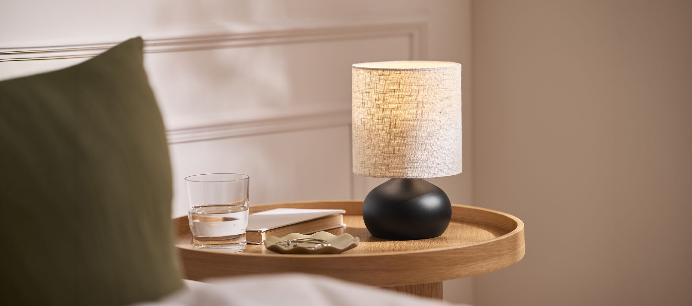 Nachttischlampe mit Stoffschirm, Wasserglas und Buch auf einem Holztablett.